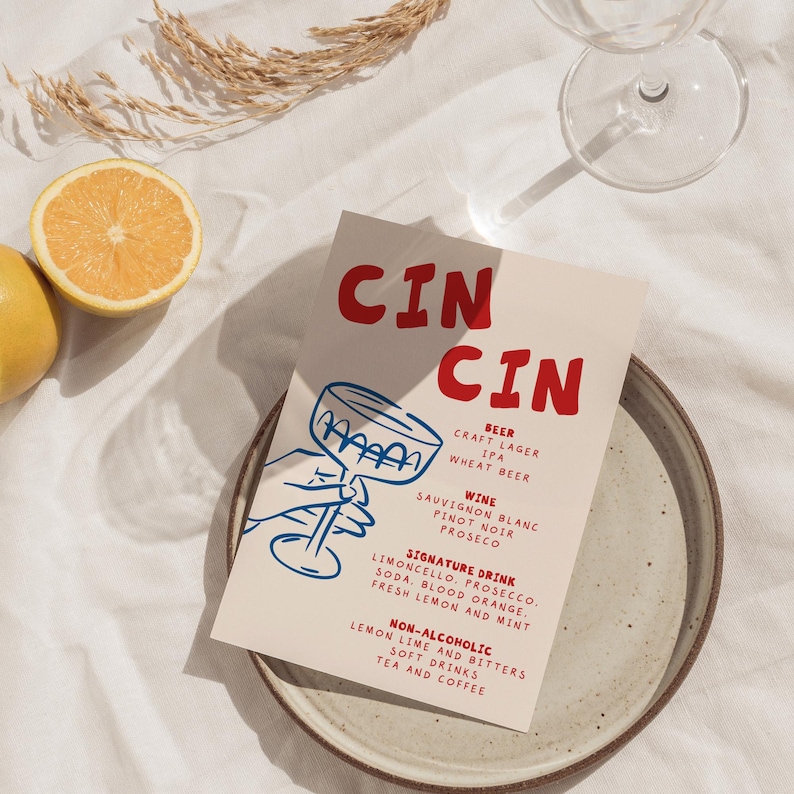 May include: A menu card with the word "CIN CIN" in red, featuring a blue illustration of a hand holding a cocktail glass. The menu lists beer, wine, signature drinks, and non-alcoholic options. A halved orange and a glass are also visible.