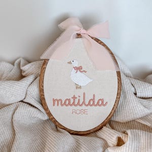 May include: An oval wooden embroidery hoop containing cream-coloured fabric with an embroidered white goose wearing a pink bow. Below the goose, the name "matilda" is stitched in pink, with "ROSE" in smaller letters. A light pink ribbon is tied at the top.