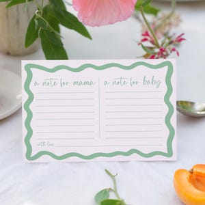 A note card with a green border and a blank space for writing is placed on a table, surrounded by a vase of flowers and a partially peeled orange.