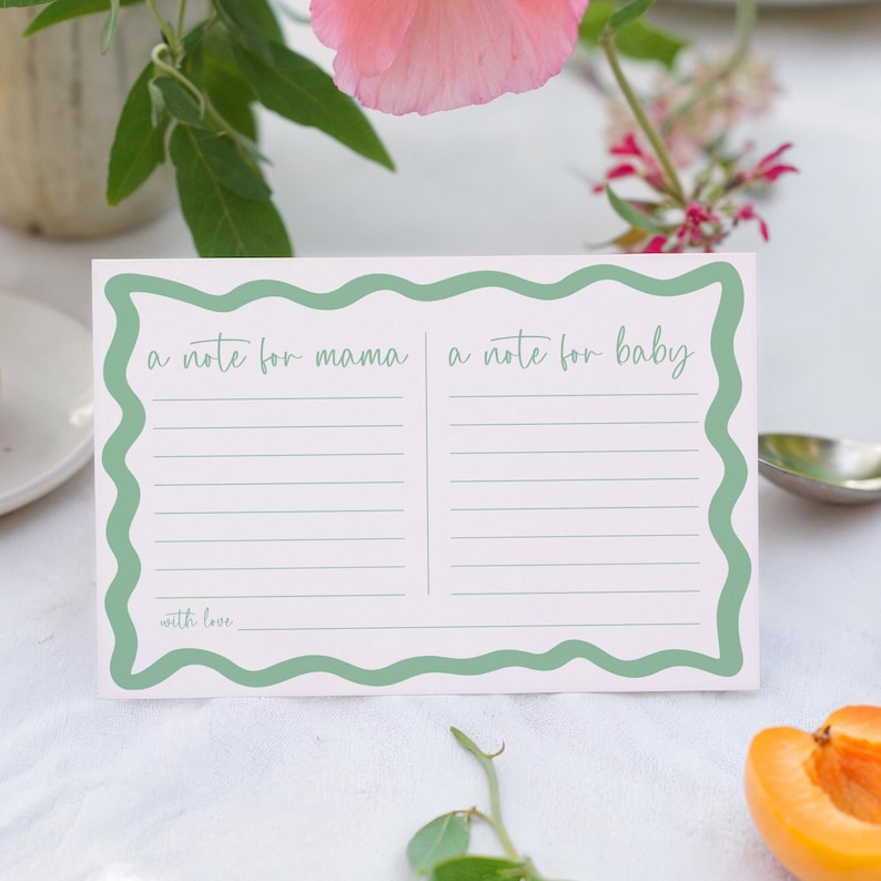 A note card with a green border and a blank space for writing is placed on a table, surrounded by a vase of flowers and a partially peeled orange.