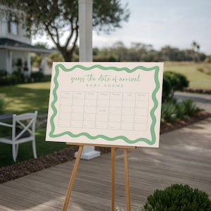 a white sign with a green border, displaying a calendar for the date of arrival of a baby named "Baby Adams". The sign is placed on a wooden deck, with a white chair and a tree visible in the background.