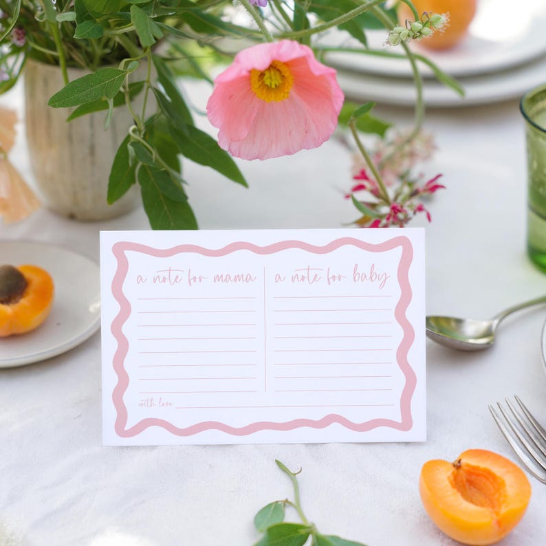 a pink flower, a note card, and some fruit on a table, with a vase of flowers in the background.