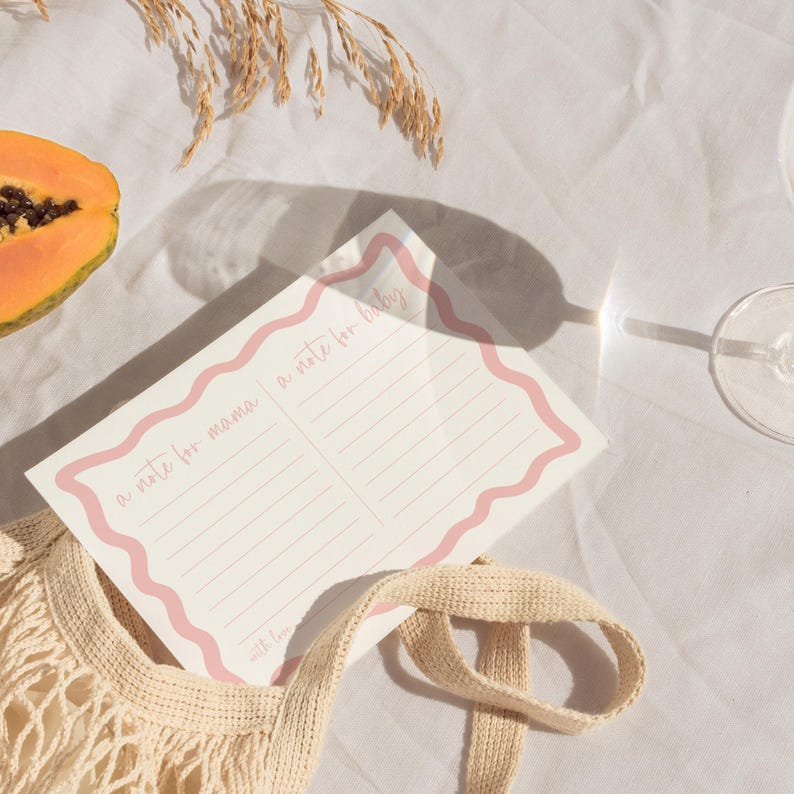 a pink and white note card with a scalloped edge, placed on a white cloth, along with a slice of papaya, a glass of wine, and a woven bag.