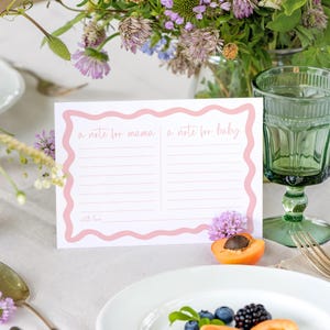 a table setting with a pink card that reads "a note for mama a note for baby with love", surrounded by various fruits, flowers, and a green glass.