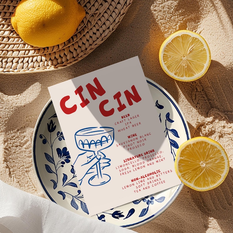 May include: A menu card with the word "CIN CIN" in red, listing beer, wine, signature drinks, and non-alcoholic beverages. The card is on a blue and white floral plate, with lemon halves and a whole lemon nearby.