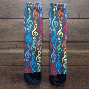 May include: A pair of crew-length socks featuring a dark gray base color with a repeating pattern of colorful treble clef musical symbols in shades of red, orange, yellow, green, and blue. The toe and heel sections are solid black in color.