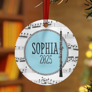 May include: Circular Christmas decoration with a musical theme. The ornament has a light blue circle with the name "SOPHIA" and the year "2025" in black text. A silver glitter border surrounds the blue circle. A black oboe is on the right, with musical notes in the background.