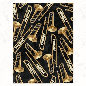 May include: A black blanket featuring a pattern of gold-colored trombones and the name "JOSHUA" in white. The trombones are arranged in various orientations across the surface, creating a musical theme.