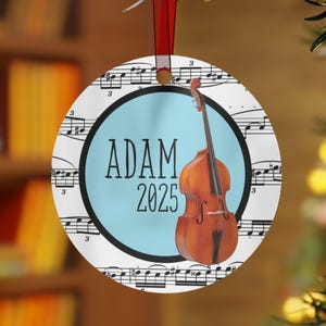 May include: Round Christmas ornament with a light blue center featuring the name "ADAM" and the year "2025". A brown double bass is on the right. The background has musical notes and a red ribbon for hanging.