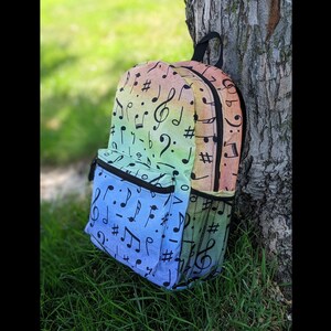 May include: A rainbow-colored backpack with black musical notes printed on it. The backpack has a zippered front pocket and a mesh pocket on the side.