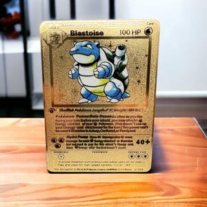 May include: A gold-colored Blastoise Pokémon card. The card features a blue and white Blastoise illustration, with text including "Blastoise," "100 HP," and attack details. The card is a Stage 2 evolution.