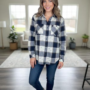 May include: A button-down plaid shirt in navy blue, white, and tan. The shirt has two chest pockets and small floral accents. The person is wearing dark blue jeans and white sneakers.