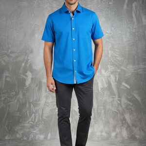 May include: A man wearing a bright blue short-sleeved button-down shirt with a chest pocket, paired with dark gray pants and brown leather shoes. The shirt has a white collar and is buttoned up. The background is a textured gray.