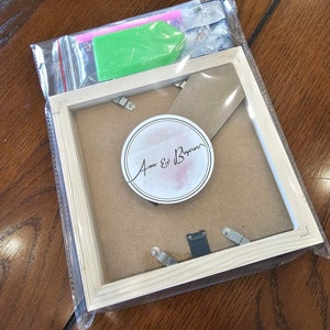 May include: A wooden picture frame with a clear glass front and a white background. The frame has a small circle sticker with the text "Ava & Bryan" in black cursive writing.