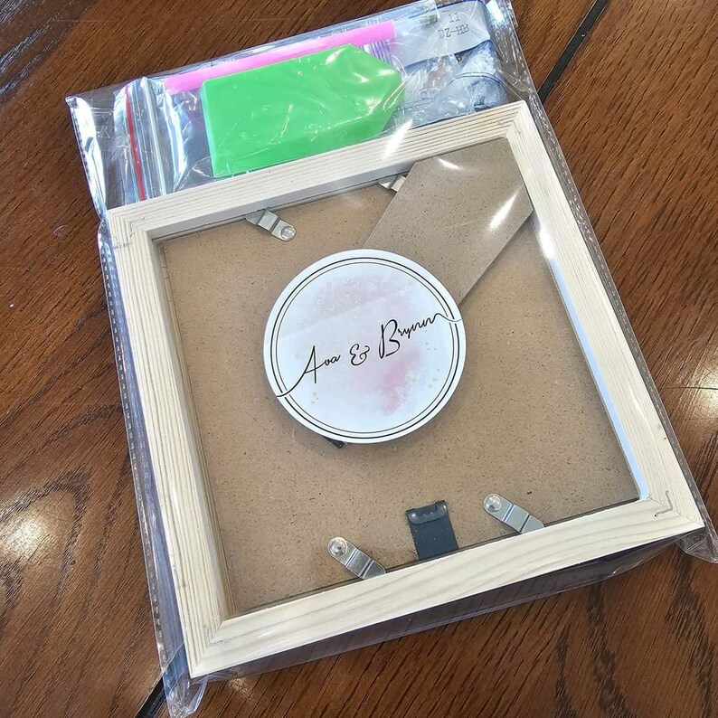 May include: A wooden picture frame with a clear glass front and a white background. The frame has a small circle sticker with the text "Ava & Bryan" in black cursive writing.