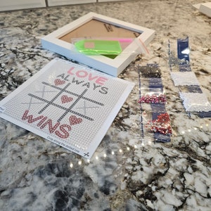 May include: A white canvas with a diamond painting design that says "Love Always Wins" with pink hearts and a tic-tac-toe board. The canvas is surrounded by small bags of colorful diamonds.