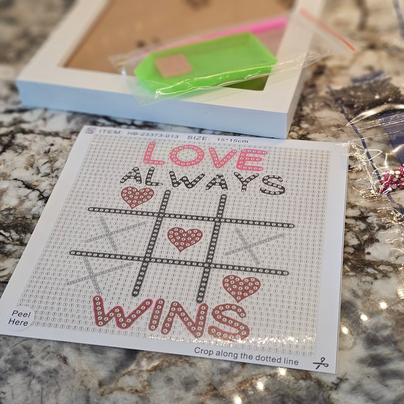 May include: A white diamond painting kit with a black and white tic-tac-toe board with pink hearts. The text "I LOVE ALWAYS WINS" is printed on the kit. The kit is 15 inches by 15 inches.