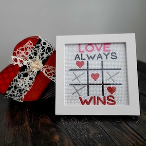 May include: White framed cross-stitch with a tic-tac-toe board and the text "Love Always Wins" in pink and red lettering. The board has red hearts in the squares.