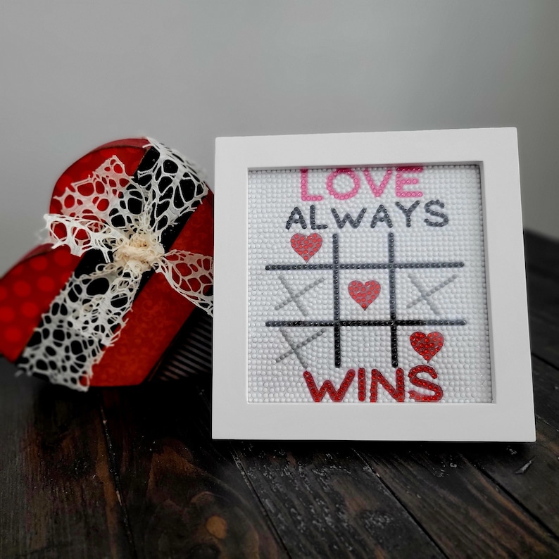 May include: White framed cross-stitch with a tic-tac-toe board and the text "Love Always Wins" in pink and red lettering. The board has red hearts in the squares.