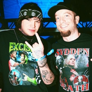 May include: Two people wearing black t-shirts with graphic prints. The person on the left is wearing a black bandana and has a tattoo on their arm. The person on the right is wearing a black baseball cap and has a beard. The t-shirt on the left says "Excision" and the t-shirt on the right says "Sudden Death".