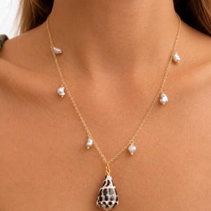 May include: A gold chain necklace featuring a central pendant made of a cone-shaped seashell with a black and white spotted pattern, accented by six small, irregular white pearls spaced evenly along the delicate gold-toned metal links.