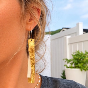 May include: Gold-toned earrings with a textured, rectangular design and a cream-colored accent. The earrings hang from a simple gold hoop. A delicate gold necklace with small pearls is also visible.