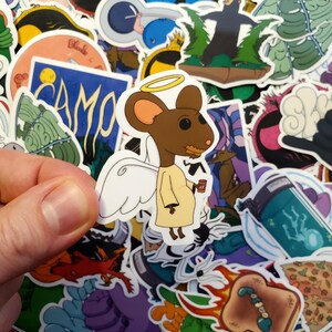 May include: A sticker of a cartoon mouse wearing a halo and wings, holding a cup of coffee. The mouse is brown with a white halo and wings. The sticker is surrounded by other stickers with various designs.