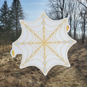 May include: A white sticker with a gold spiderweb design. The web is intricate and detailed, with a radial pattern. The sticker is shaped like a star.