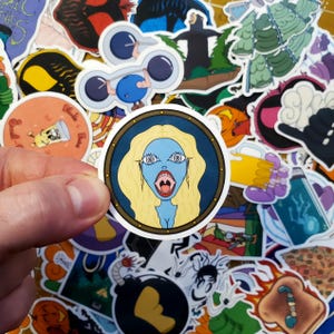 May include: A sticker featuring a woman with blue skin and wide, staring eyes. She has long blonde hair and is wearing a blue dress. The sticker is round and has a gold border.