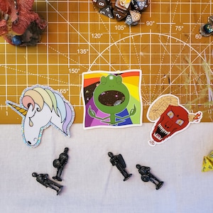 May include: A set of four stickers featuring a green frog meditating, a unicorn with a rainbow mane, a red devil with horns, and a black and white illustration of a knight. The stickers are on a white surface with a yellow cutting mat in the background.