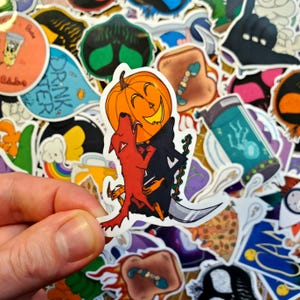 May include: A sticker featuring a cartoon pumpkin with a toothy grin, wearing a black hat and holding a red dog. The pumpkin is holding the dog's head in its mouth. The sticker is on a white background.