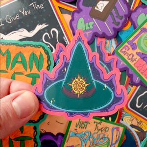 May include: A sticker featuring a green witch hat with a gold sunburst design, surrounded by a pink and purple border. The sticker is held in a hand, with other colorful stickers in the background. The sticker has a mystical theme.