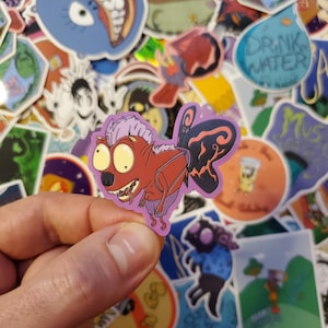 May include: A sticker of a cartoon creature with red skin, purple hair, and black wings. The creature has large eyes and a wide mouth. The sticker is on a purple background.