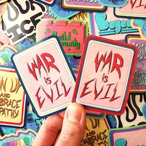 Könnte beinhalten: Zwei Aufkleber mit dem Text "WAR is EVIL" in roter, stilisierter Schrift auf rosa Hintergrund. Die Aufkleber haben einen blauen und roten Rand. Weitere bunte Aufkleber befinden sich im Hintergrund.