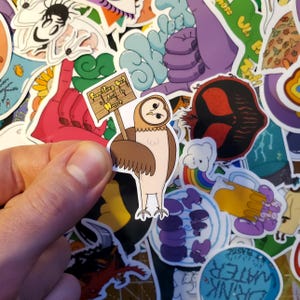 May include: A brown and white owl sticker holding a sign that says "Something Bad is About to Happen". The sticker is on a background of other colourful stickers.