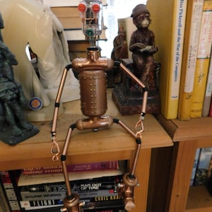 May include: A handmade copper robot sculpture with a cylindrical body, articulated limbs, and a head with red and green lights. The robot is positioned on a wooden shelf, surrounded by books and other decorative items.