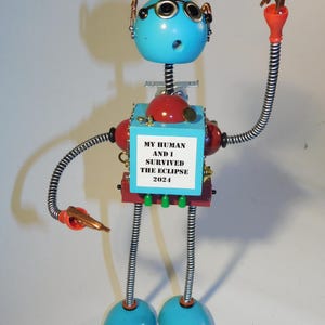 Puede incluir: Una escultura de robot caprichosa con cabeza y pies turquesa, cuerpo rojo y brazos flexibles. El robot tiene gafas redondas pequeñas y un letrero que dice "MY HUMAN AND I SURVIVED THE ECLIPSE 2024."