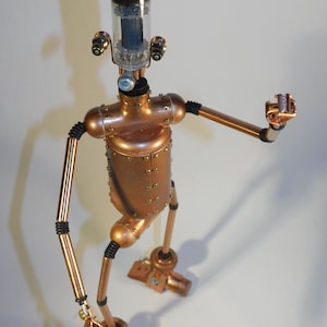 May include: A copper-toned steampunk robot sculpture with a cylindrical body and articulated limbs. The robot has a glass tube head and small, round eyes. The arms are extended, and the feet are rectangular. The robot is made of metal and has a vintage aesthetic.