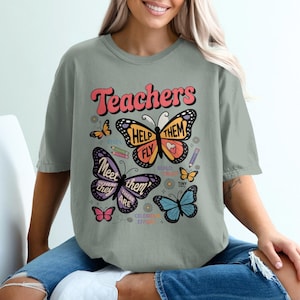 May include: Sage green t-shirt featuring a butterfly design. The graphic includes the word "Teachers" in a retro font, along with butterflies and the phrases "Help Them Fly" and "Meet Them Where They Are."