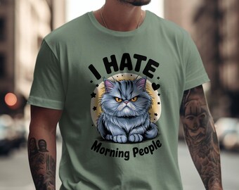 May include: Sage green t-shirt featuring a grumpy cartoon cat with the text "I HATE Morning People." The cat is gray with yellow eyes, set against a yellow circle with stars and hearts. A humorous graphic tee.