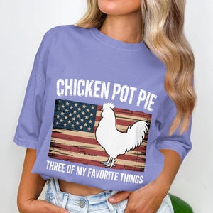 May include: A light purple t-shirt with the text "CHICKEN POT PIE" above an American flag graphic and a white chicken illustration. Below the graphic, the text reads "THREE OF MY FAVORITE THINGS."