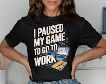 May include: Black t-shirt with the text "I PAUSED MY GAME TO GO TO WORK" in white and yellow. The design includes a blue game controller, a laptop, and a notepad with a pencil. The shirt is a casual wear item.