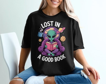 May include: Black t-shirt with a graphic of a green alien reading a book, seated in a purple chair. The shirt has the text "LOST IN A GOOD BOOK" in white. A mug, planets, and a cookie are also in the graphic.