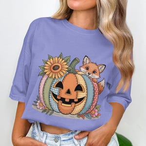 May include: A lilac t-shirt with a colourful pumpkin design. The pumpkin has a jack-o'-lantern face, a sunflower, and a fox. The pumpkin is decorated with patchwork patterns. The shirt is a comfortable, casual style.
