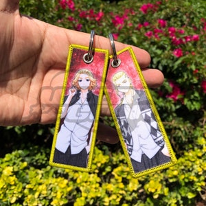 以下が含まれることがあります： アニメキャラクターのイラストが描かれた長方形のキーホルダー2つ。各キーホルダーは、キャラクターの画像が入った赤い背景、黄色の縁取り、シルバーのキーリングが特徴です。キャラクターは黒いジャケットと白いシャツを着ています。
