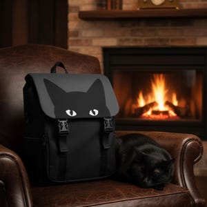 Może przedstawiać: Czarny plecak z motywem kociej twarzy na klapie, z czarną sylwetką kota z białymi oczami. Plecak ma regulowane paski i klamry. Czarny kot zwinięty na skórzanym krześle.