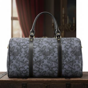 May include: A gray and black patterned duffel bag with black leather handles and accents. The bag features a floral design and gold-colored hardware. The bag is sitting on a wooden surface.