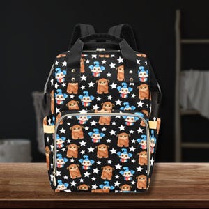 May include: Black diaper bag with a repeating pattern of brown cartoon animals and blue cartoon characters with white stars. The bag has a top handle, adjustable shoulder straps, and multiple pockets for storage.