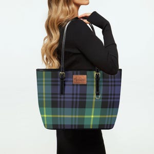 May include: A tote bag featuring a tartan design in blue, green, and yellow. The bag has black handles and trim, with gold-coloured hardware. A small, rectangular leather patch with the name "Jessica" is attached to the front.