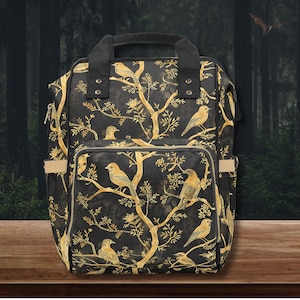 Op de afbeelding: Zwarte rugzak met een gouden boom- en vogelpatroon. De rugzak heeft een handvat aan de bovenkant, een voorvak en zijvakken. Het ontwerp toont vogels op takken, wat een natuurlijke uitstraling geeft.